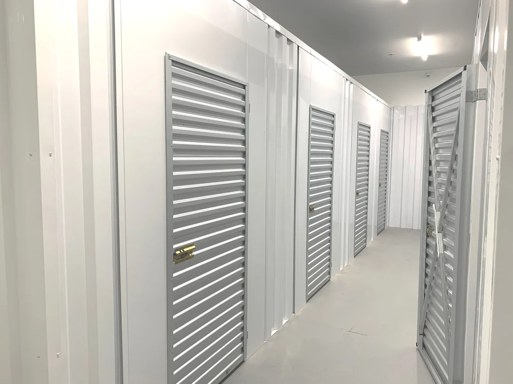 Interior hallway of climate-controlled storage units with clean white walls, gray metal roll-up doors, and bright overhead lighting.
