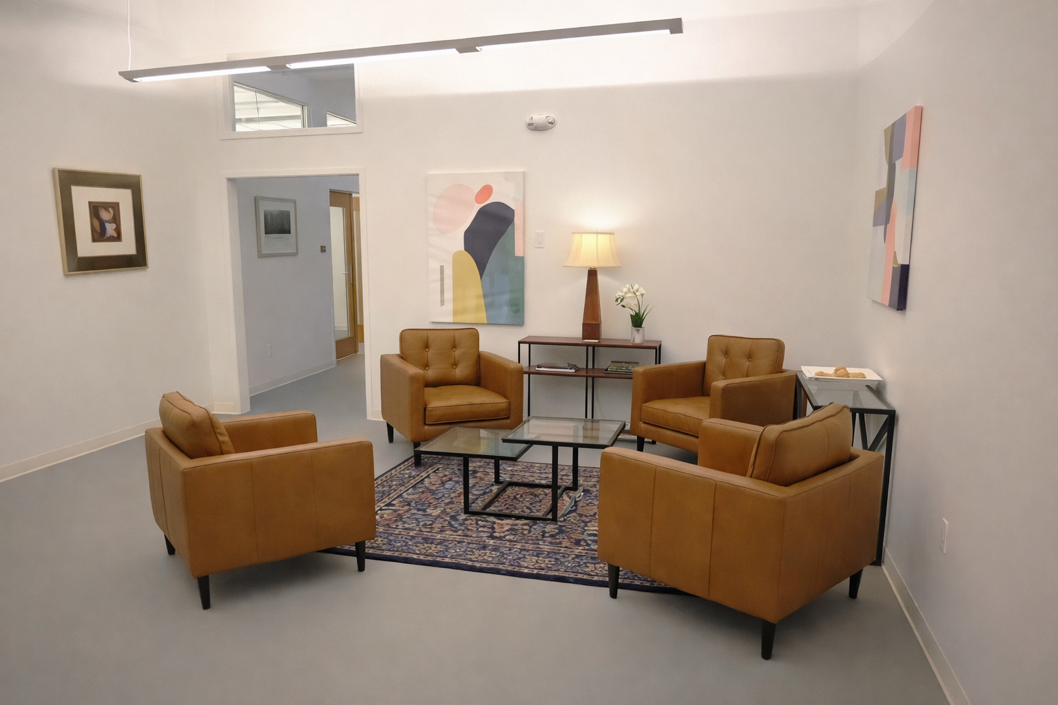 Modern commercial lobby lounge with four caramel leather armchairs around a glass coffee table on a patterned rug, accented by abstract wall art and a console table with a lamp.