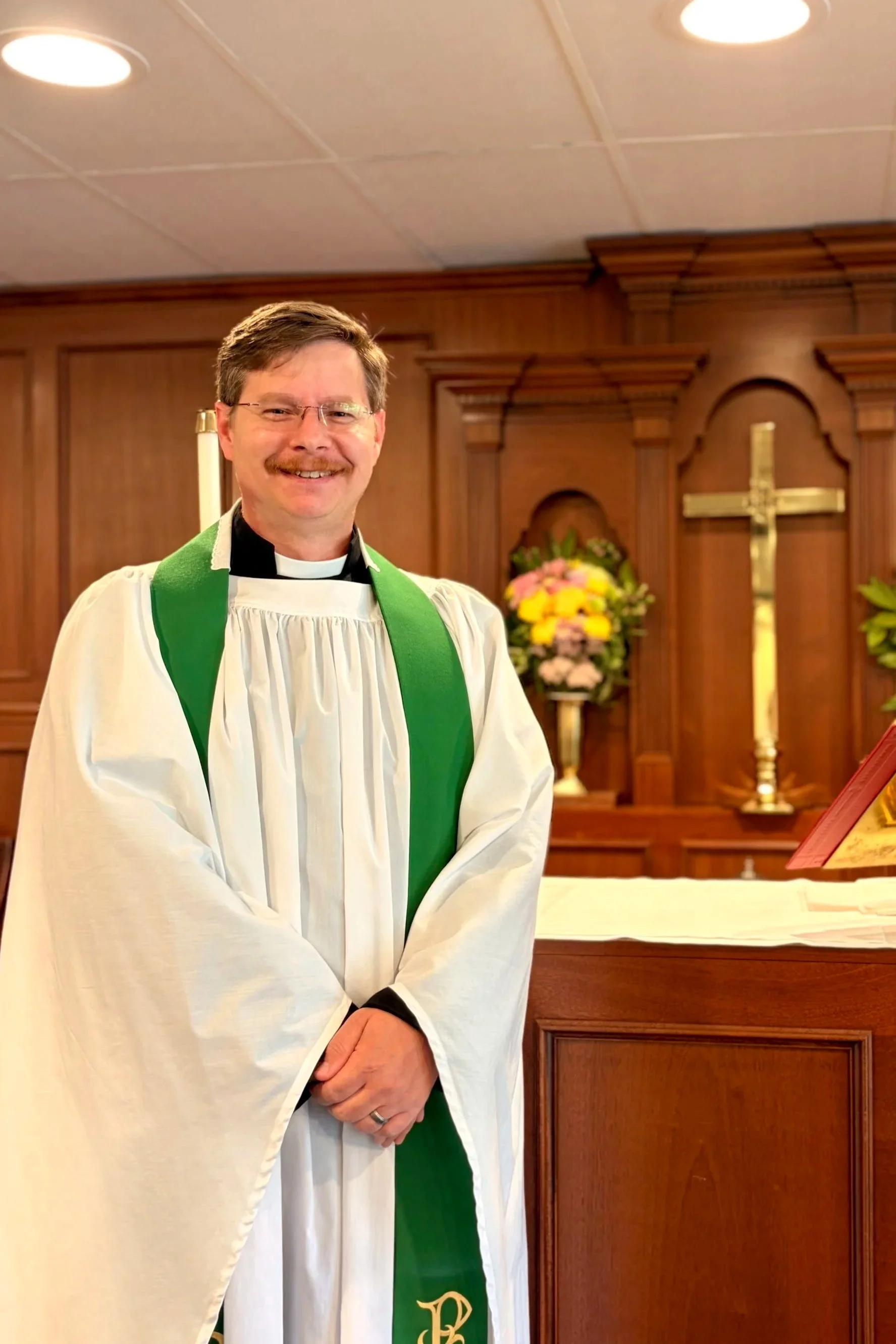 A priest in white robes with a green sash standing in front of an altar with a cross and floral arrangements.