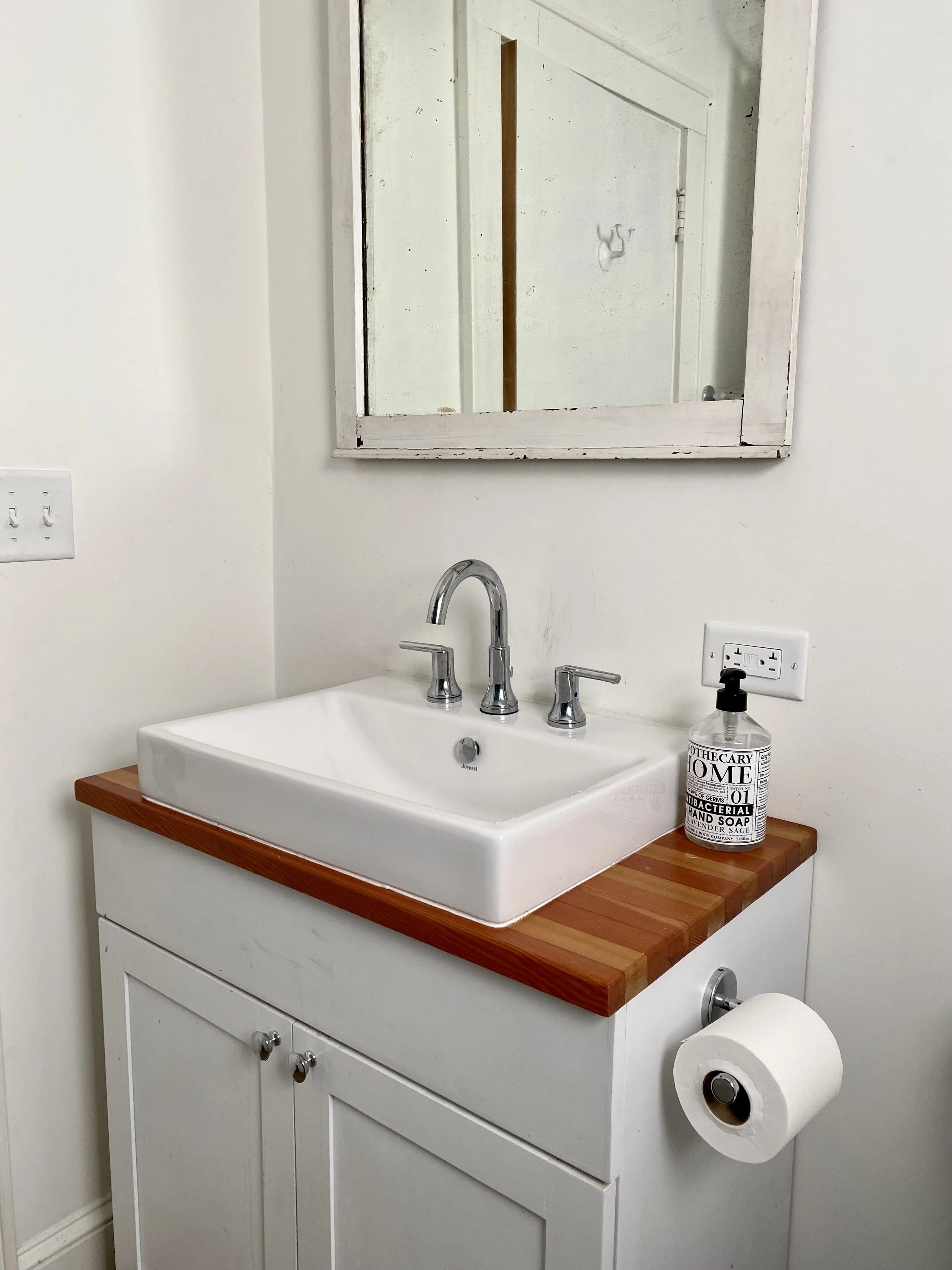 Bathroom sink with a wooden countertop, a rectangular mirror, silver faucet, hand soap bottle, and toilet paper roll.