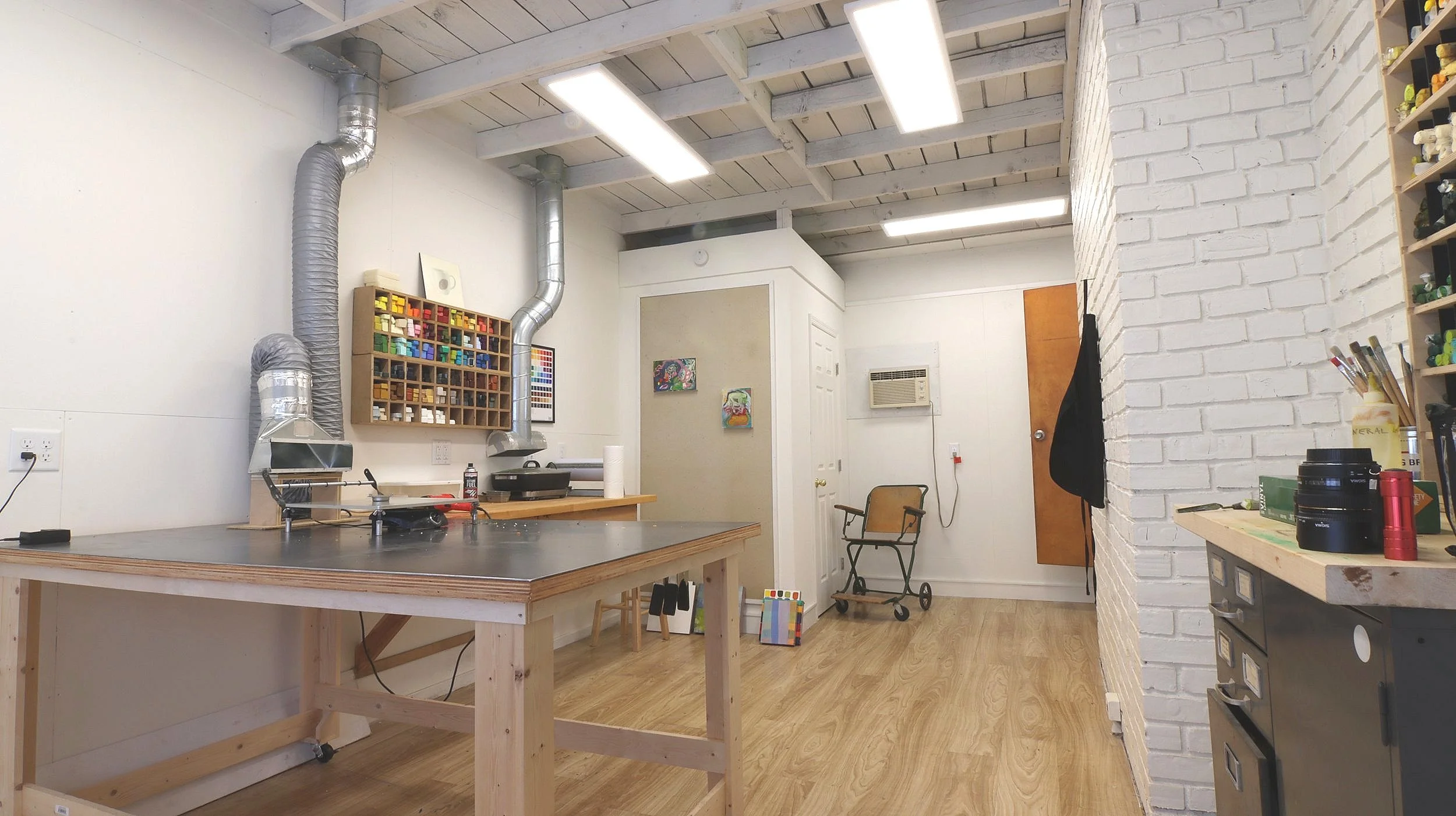 Workshop interior with wooden workbench, shelves with paint, ventilation ducts, air conditioner, and art supplies on a counter.