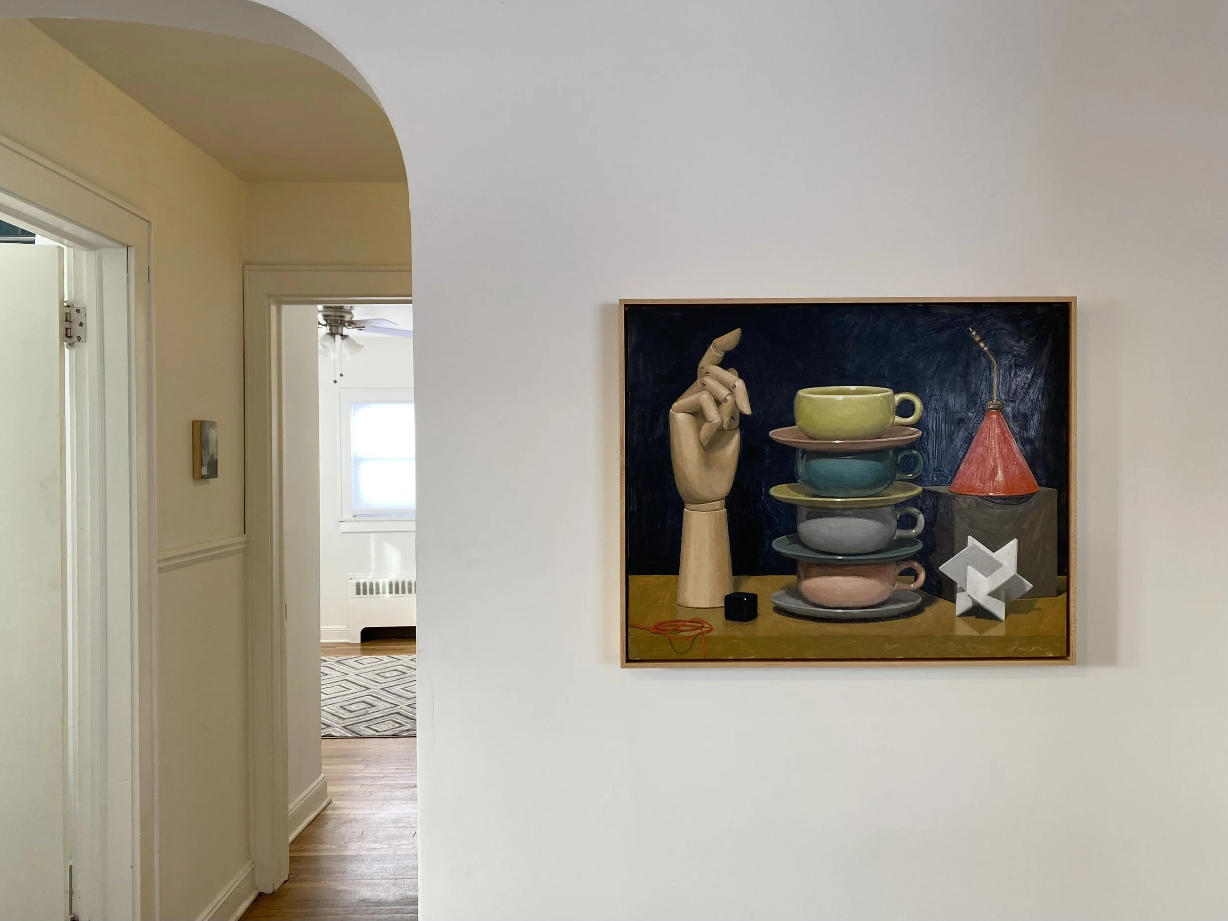 Interior of a room with a hallway, featuring a framed painting on the wall. The painting depicts a still life with stacked teacups, a wooden hand, and geometric shapes.