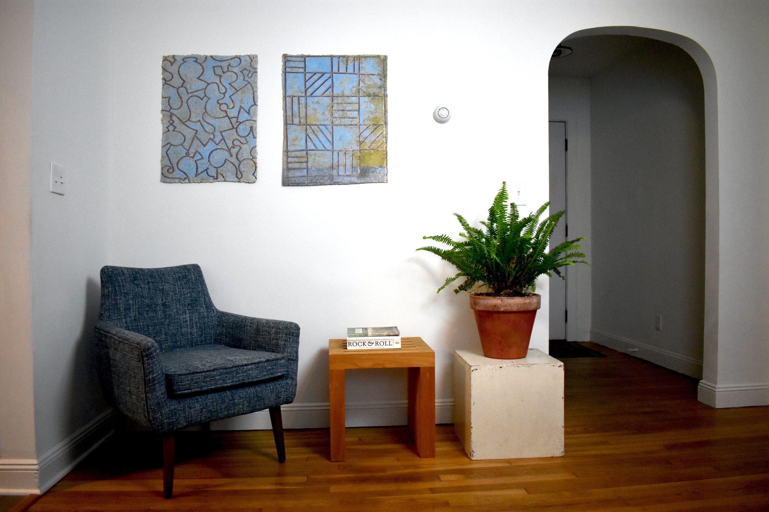 Interior of a modern living space with a blue armchair, wooden side table holding books, a potted fern on a white pedestal, and abstract art on the white walls.