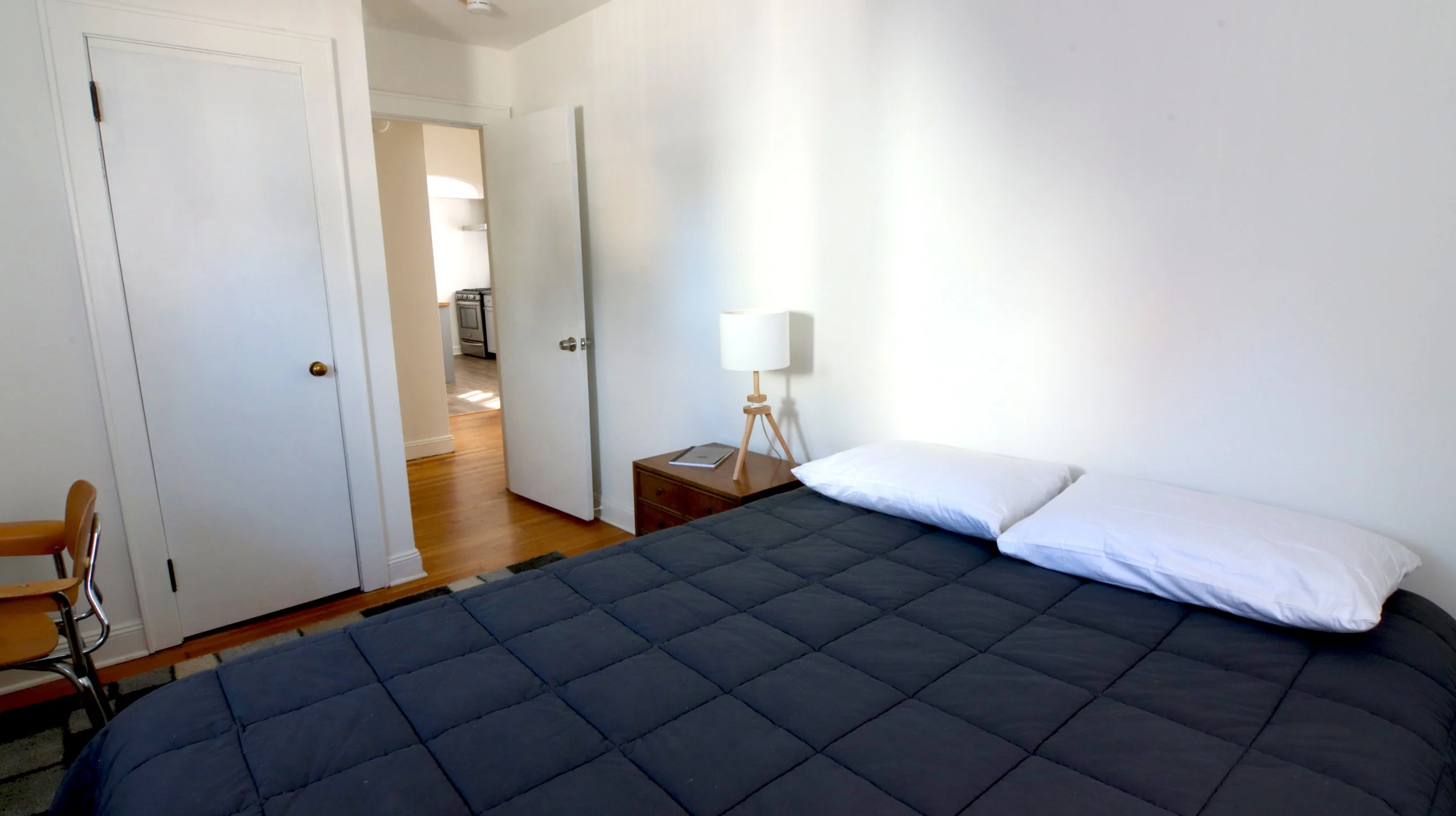 Minimalist bedroom with bed, nightstand, lamp, and open door to hallway.