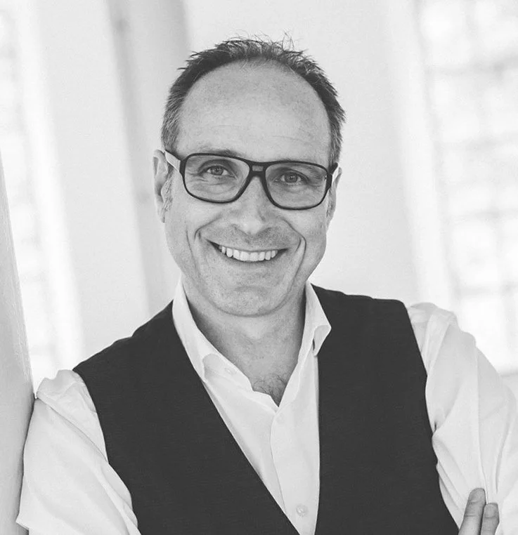 Porträt eines lächelnden Mannes mit Brille und Weste, schwarz-weiß.