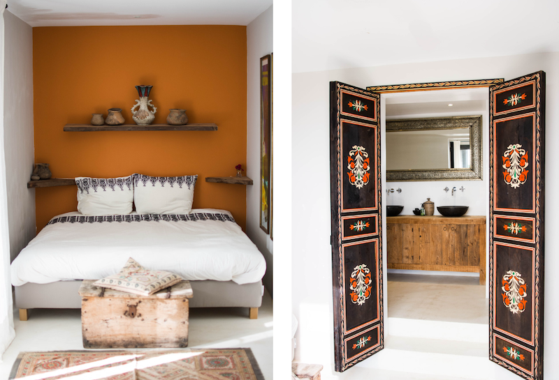 Schlafzimmer mit oranger Wand und dekorativen Regalen über dem Bett, neben einem Badezimmer mit kunstvoll verzierten Türen.
