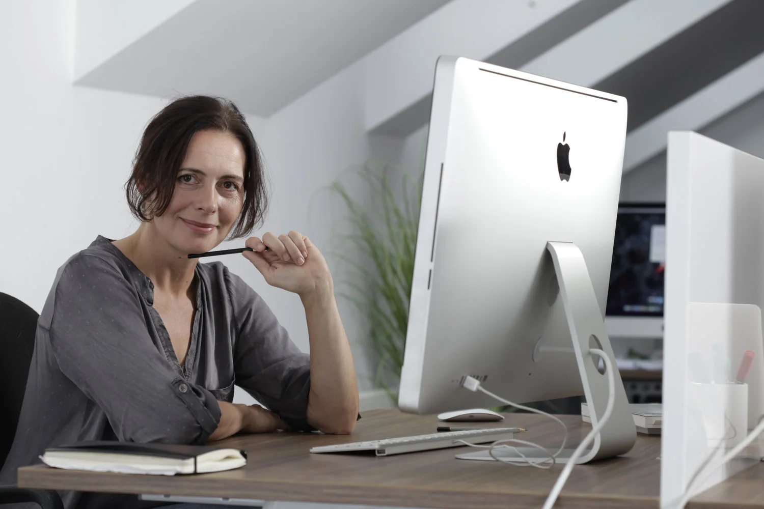 A woman sitting at a desk with an iMac computer, holding a pen near her chin and smiling at the camera.