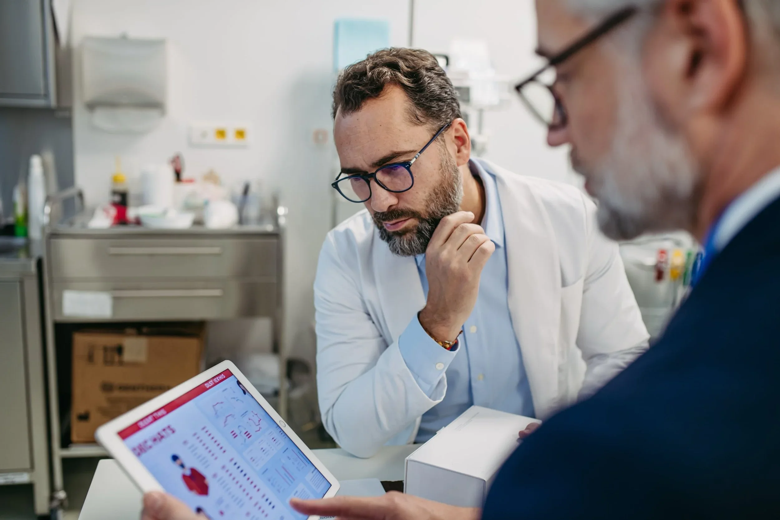 Two male doctors, one younger and one older, are discussing medical data in a hospital or clinical setting. The younger doctor is holding a tablet with Medpad PRO app, while the older looks thoughtful.