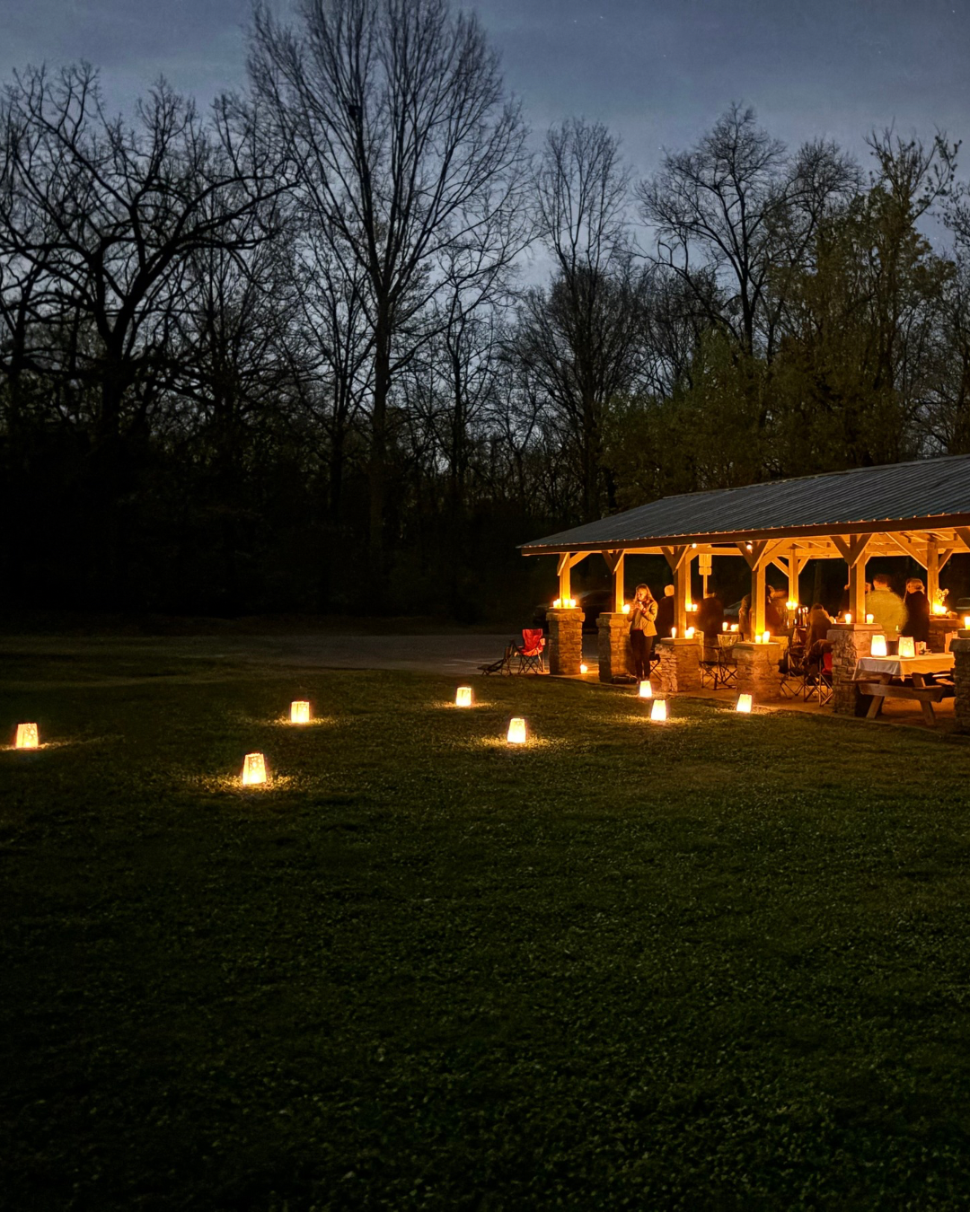 Nashville residents gathered for a community grief circle, under a lit pavilion outside at night, with glowing lanterns on the ground surrounding the area as certified Death Doula Jade Adgate guides a community event.