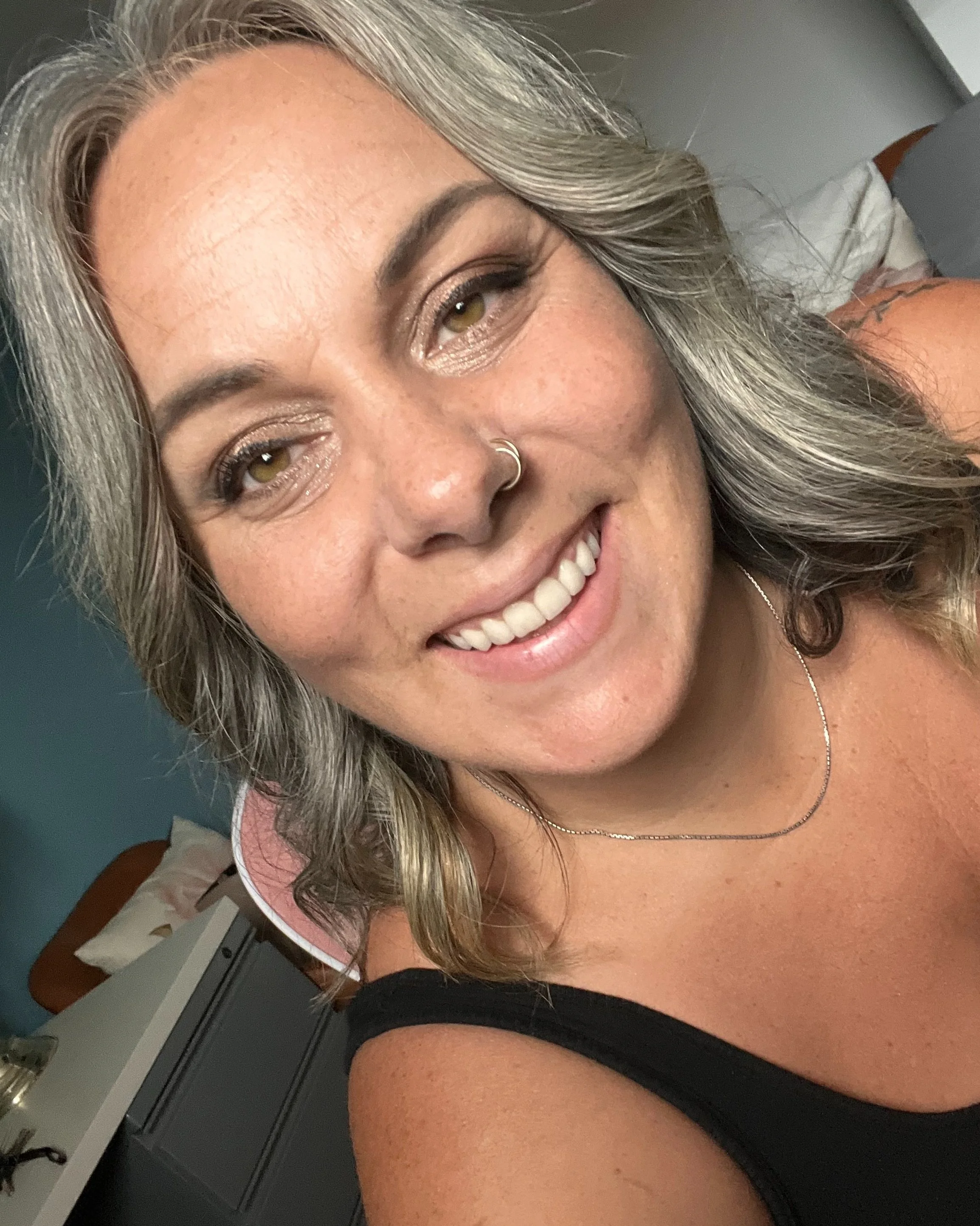 Close-up of a smiling woman with wavy, shoulder-length gray hair, wearing a black tank top and a delicate necklace, in a room with a bed and dresser in the background.