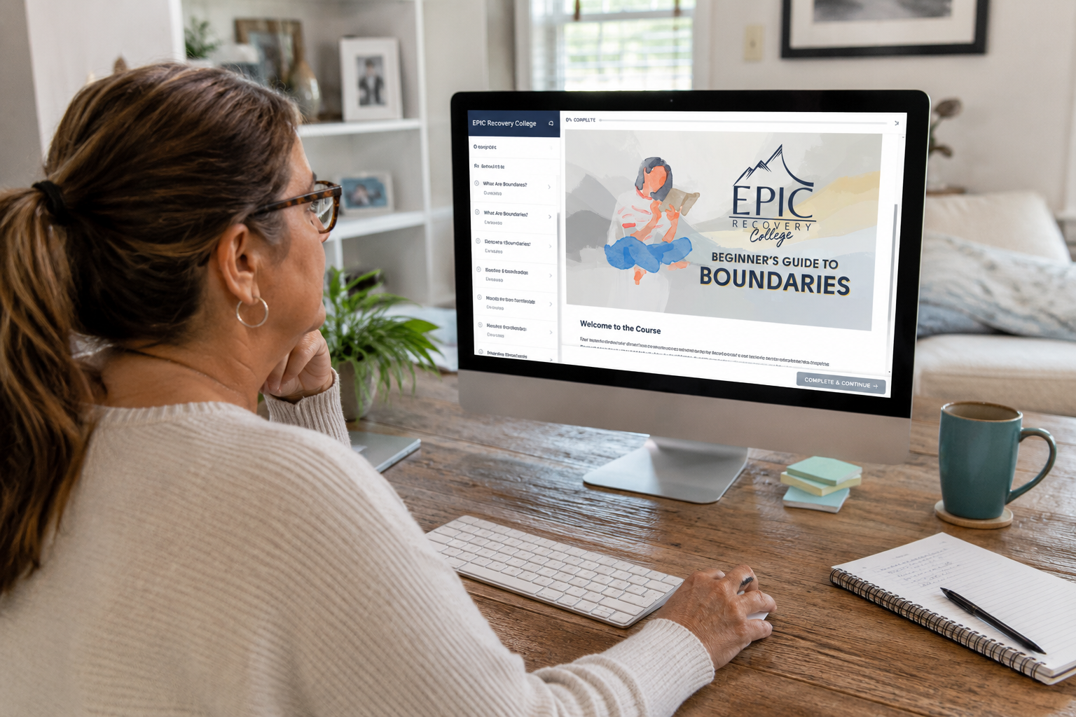 Woman sitting at a wooden desk, viewing an online course titled 'Beginner's Guide to Boundaries' on an iMac computer in a cozy, well-lit room with a bookshelf, plants, and framed pictures.