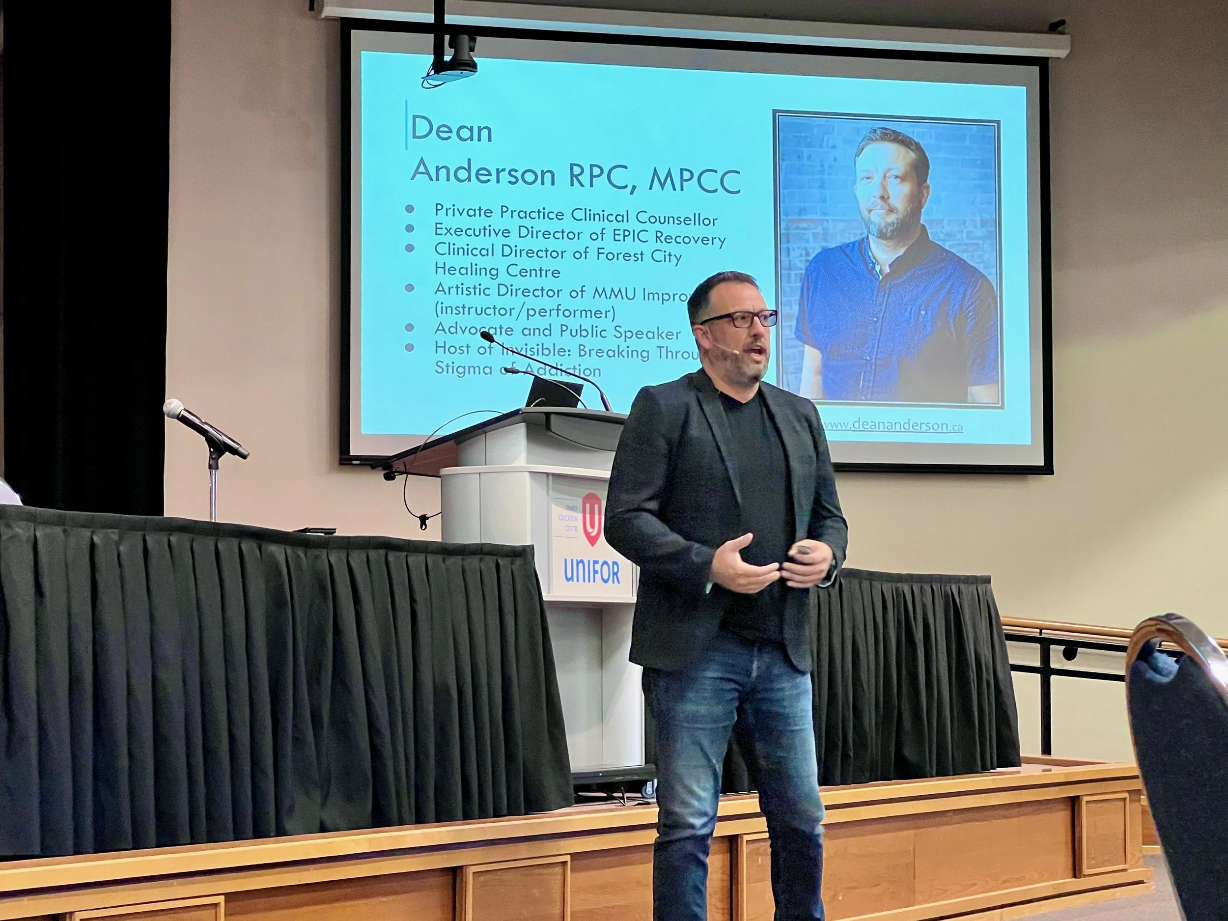 A man with glasses, wearing a dark blazer and jeans, is speaking at a presentation event, standing in front of a projection screen with a slide about Dean Anderson. The slide includes his photo and professional details.