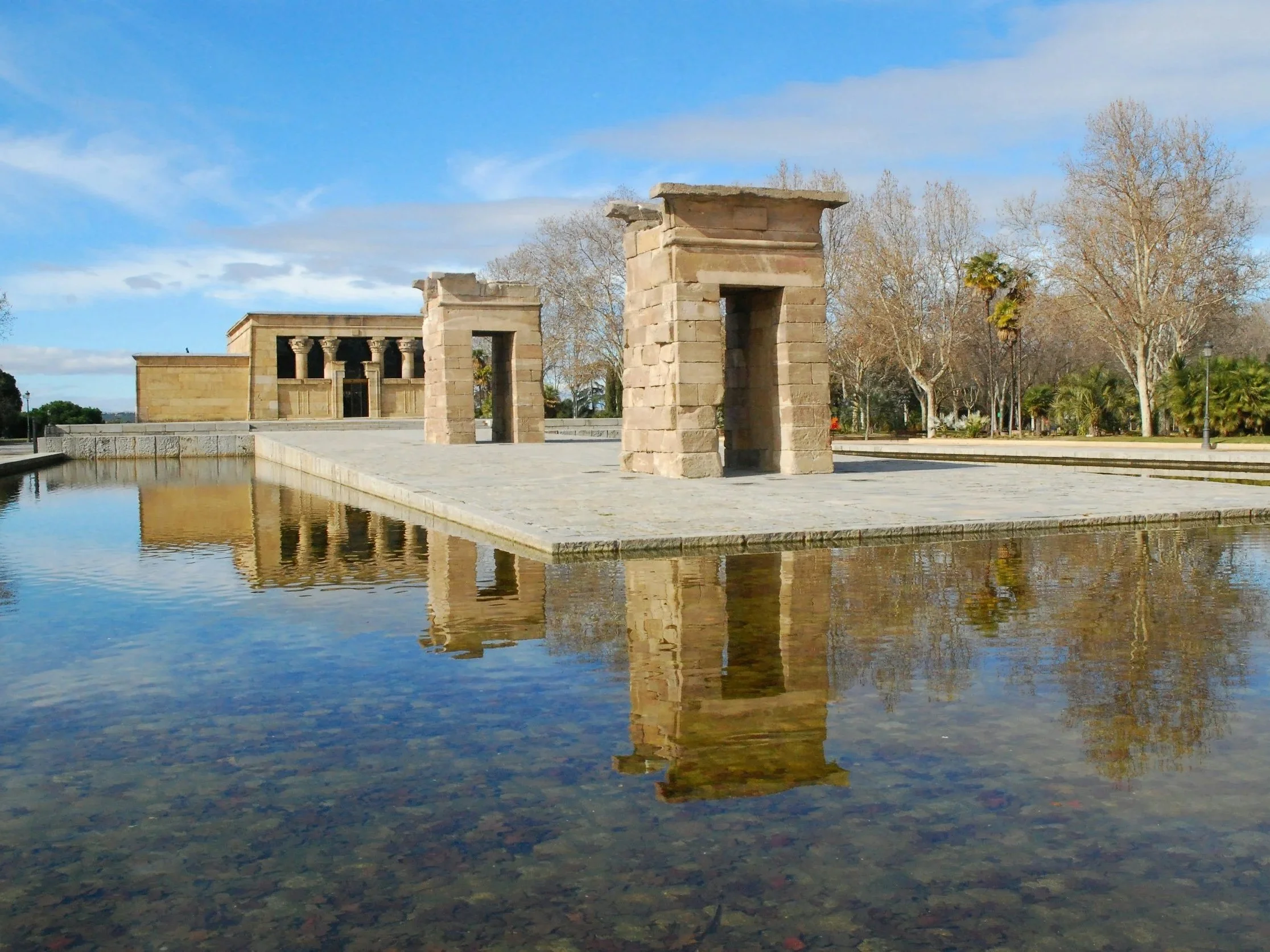 Ancient stone arch ruins next to reflective pool of water, clear sky with some clouds, leafless trees in background.