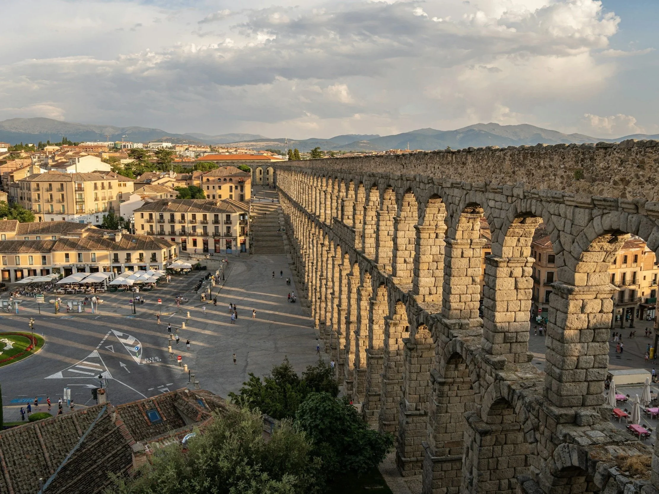 Photo of an ancient Roman aqueduct with standing arches, overlooking a city square with buildings, people, and outdoor cafes, under a partly cloudy sky with mountains in the background.