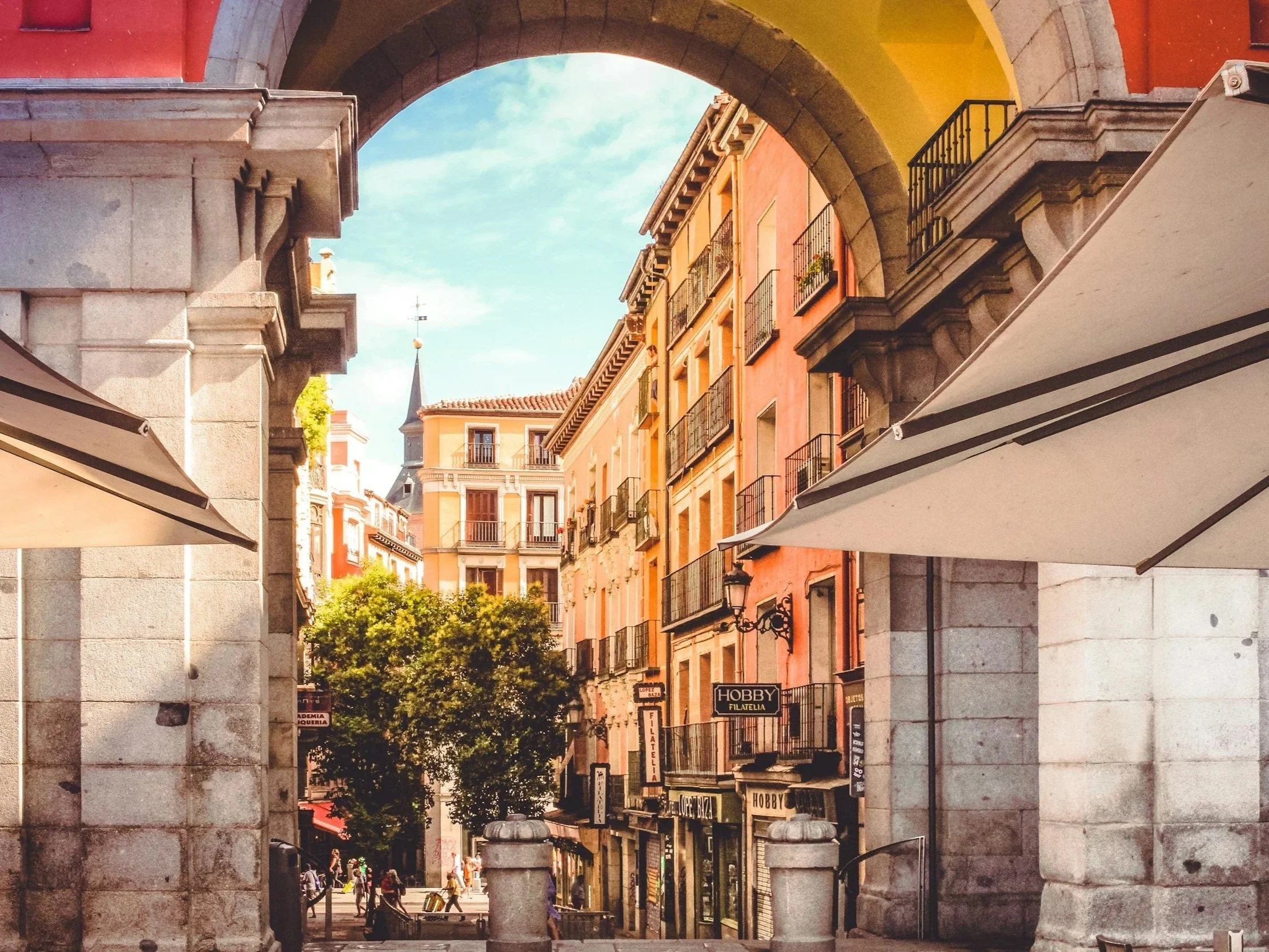 A street view of colorful buildings with balconies, seen through an archway with stone pillars. There are awnings at the bottom of the image, and trees and people walking on the street.