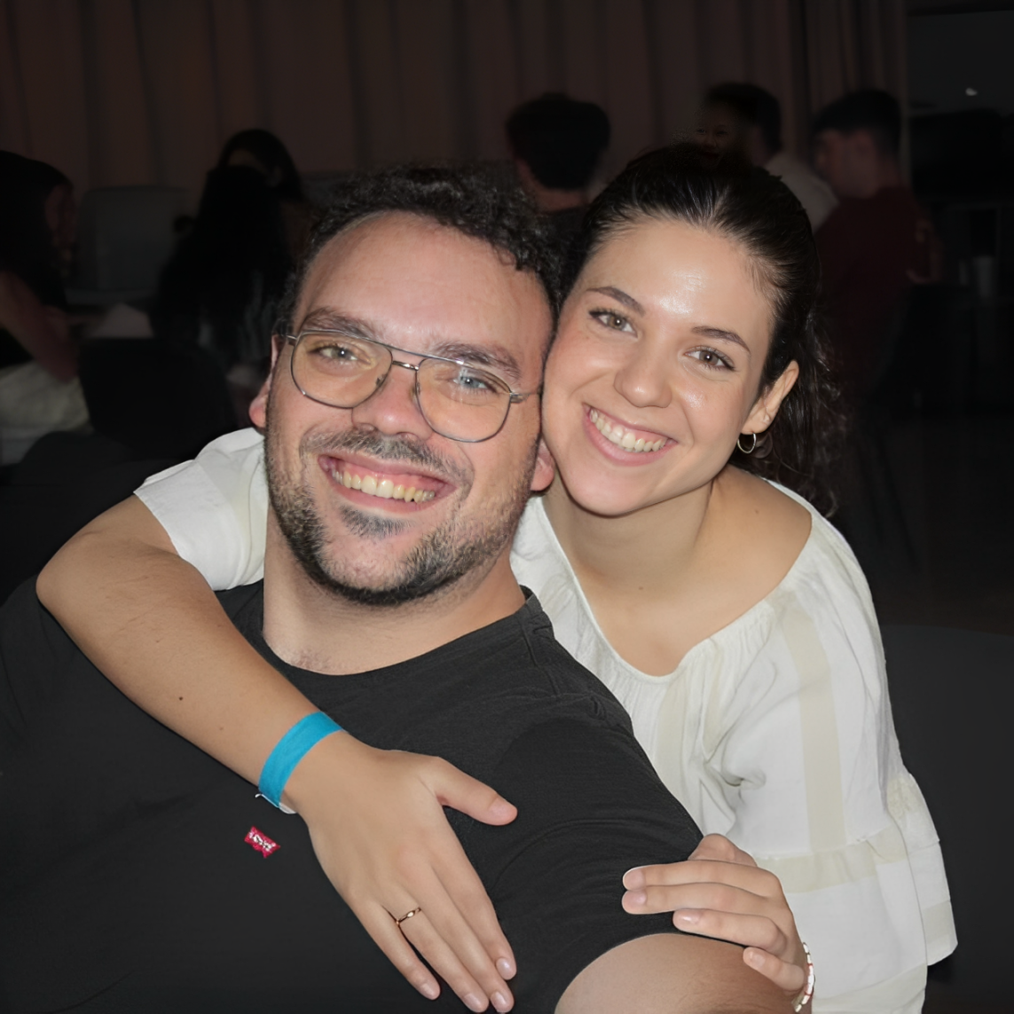Un hombre sonriente con gafas y una mujer de pelo oscuro, ambos felices, abrazándose en un lugar interior lleno de gente.