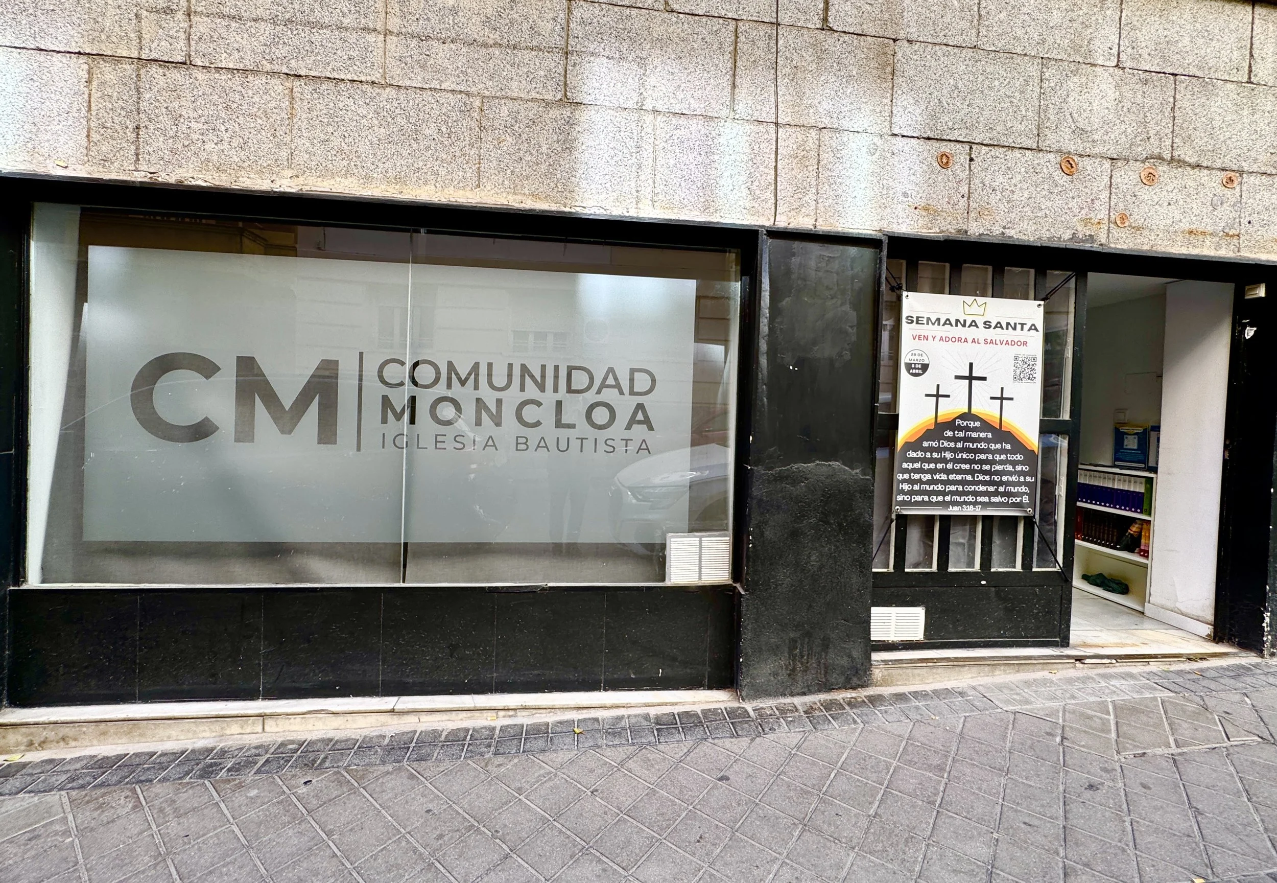 Un local con un escaparate de cristal que muestra el logotipo de Comunidad Moncloa Iglesia Bautista y un cartel adjunto con contenido religioso en español.
