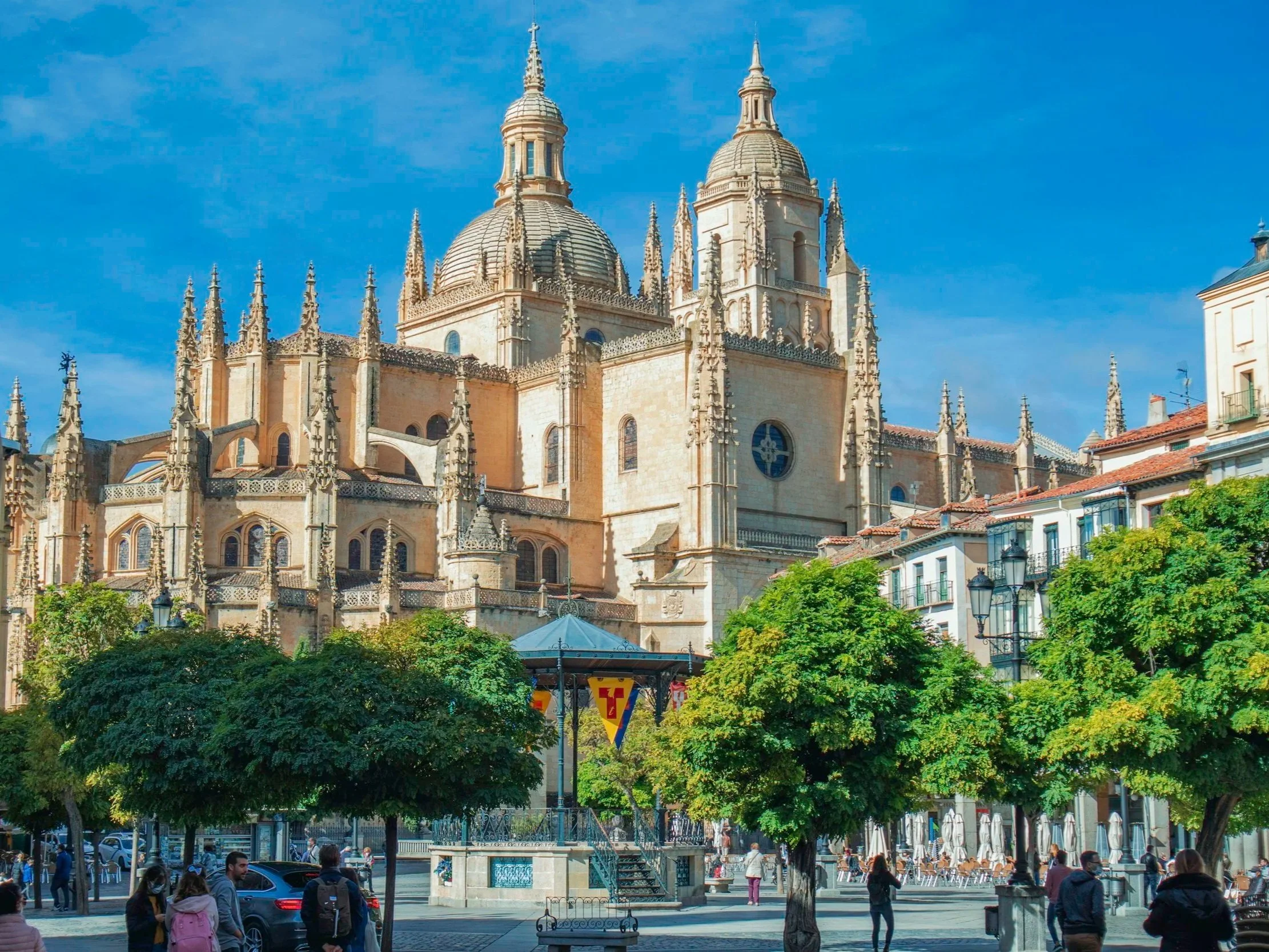 A historic cathedral with ornate Gothic architecture, large domes, and spires, situated in an urban square with trees, people, and outdoor seating.