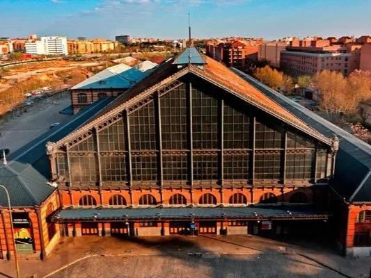 A large, industrial-style building with a triangular glass roof and brick arches at the base, set against a cityscape with modern buildings and a clear sky.