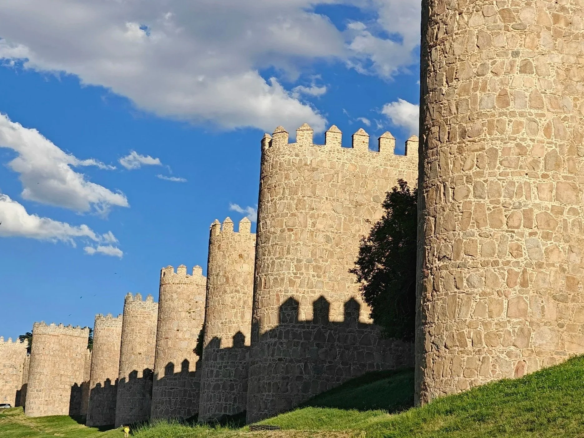Stone castle walls and towers against a blue sky with clouds, casting shadows on the ground.