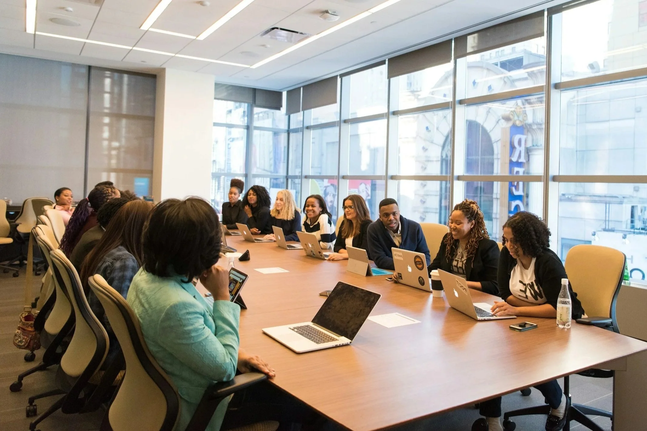 A diverse group of people sit around a large conference table in a modern office with large windows, working on laptops and talking, during a business meeting.