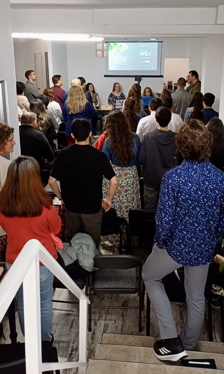 Un grupo de personas reunidas en una sala pequeña, de pie y mirando hacia una pantalla de presentación al frente, mientras algunas personas están sentadas en una mesa. La diapositiva de la presentación se ve parcialmente en la pantalla.