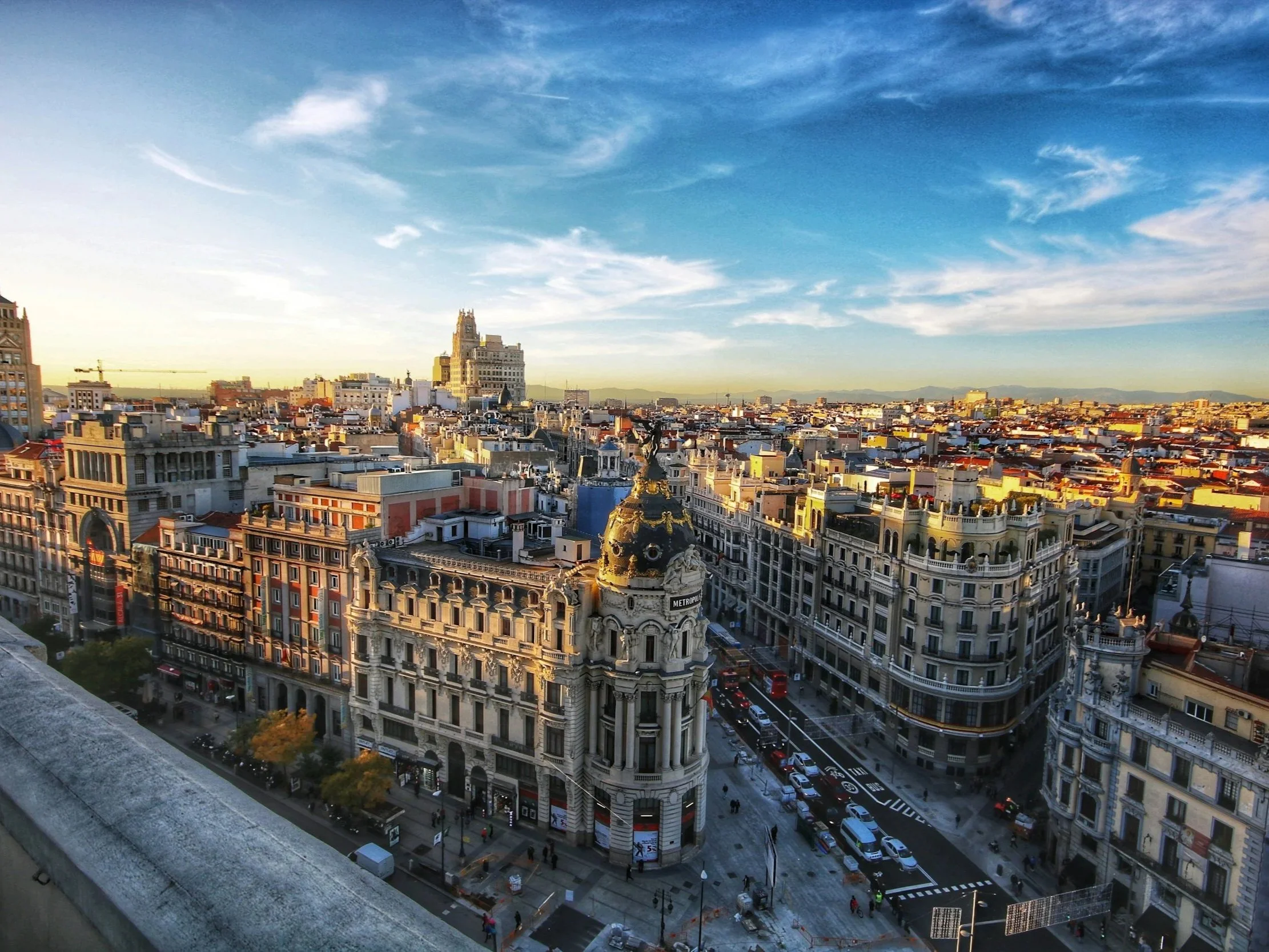 A cityscape view of downtown Madrid, Spain, with historic buildings, street traffic, and a clear sky with wispy clouds at sunset.