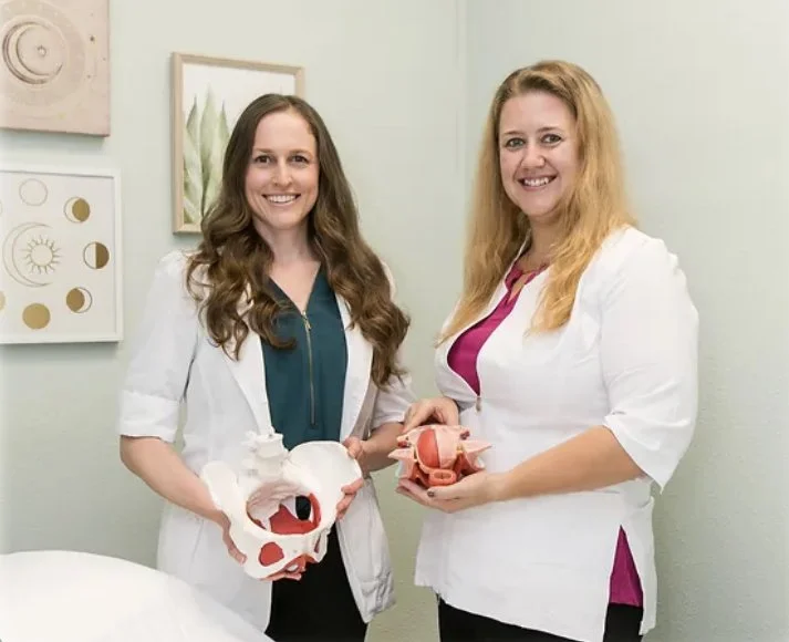 Two women standing in a room with light green walls, holding anatomical models of the human pelvis and reproductive organs, smiling at the camera.