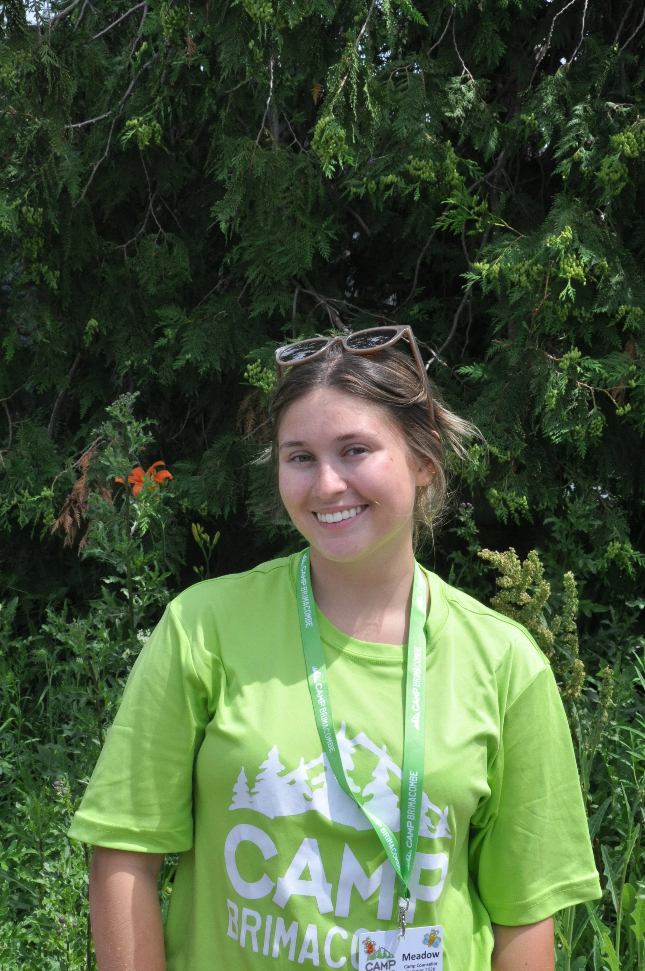 A young woman withbrown hair, wearing sunglasses on her head and a lime green T-shirt that reads 'CAMP BRIMACOMBE' in white, standing outdoors in front of lush green foliage, smiling at the camera.
