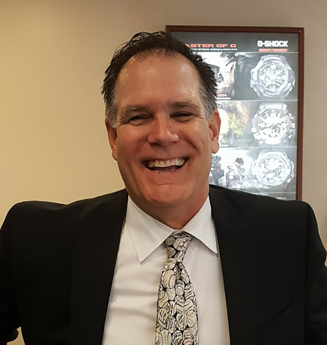 A man in a black suit, white shirt, and patterned tie smiling in an indoor setting, with a poster of G-Shock watches in the background.