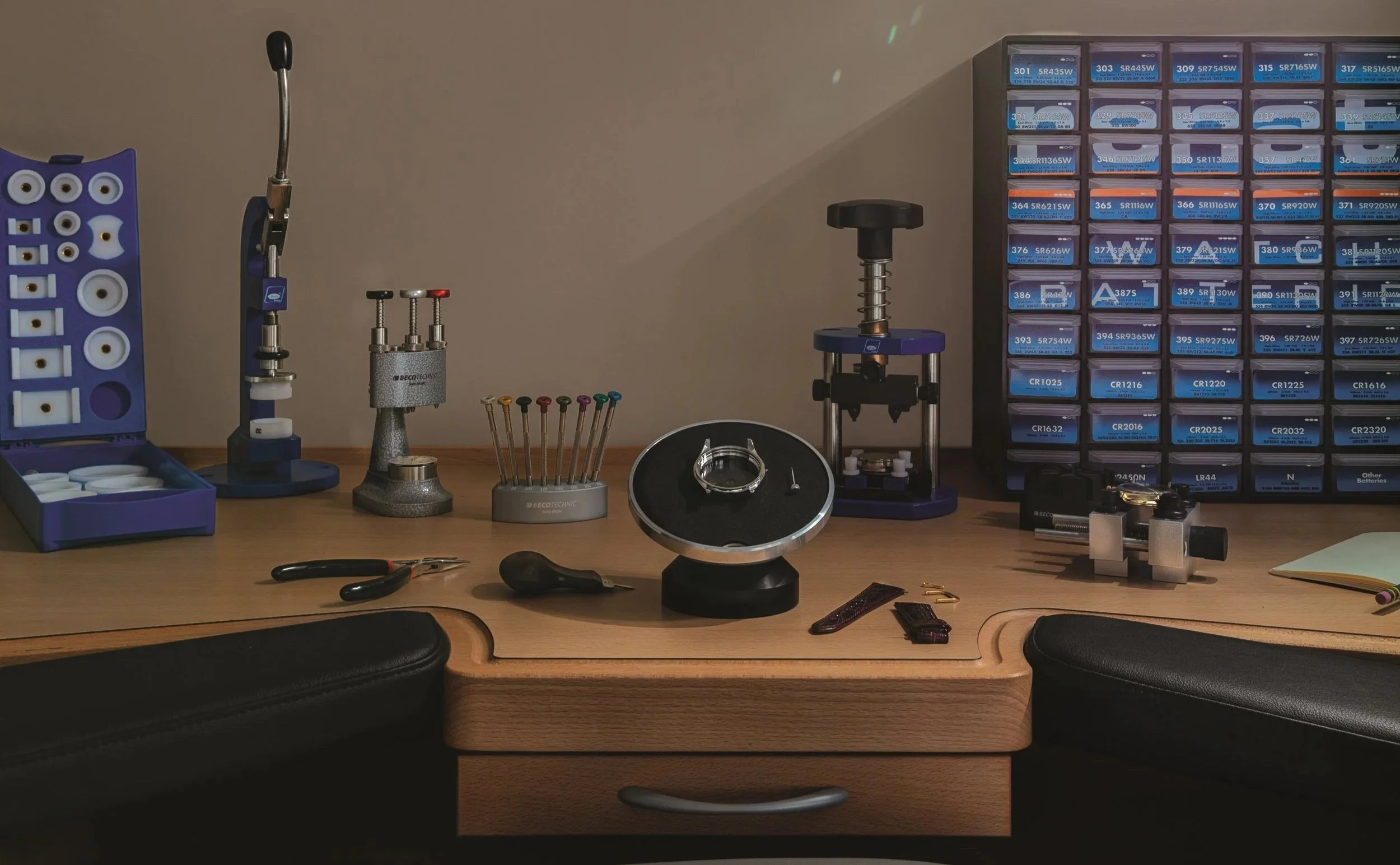 A workbench with tools, small boxes, and equipment related to watch or jewelry repair, including screwdrivers, a loupe, a soldering station, and organized storage boxes.