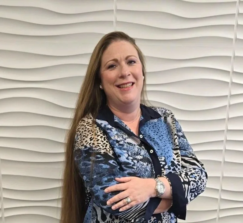 A woman with long brown hair smiling and standing with arms crossed in front of a white textured wall. She is wearing a patterned blouse and jewelry.