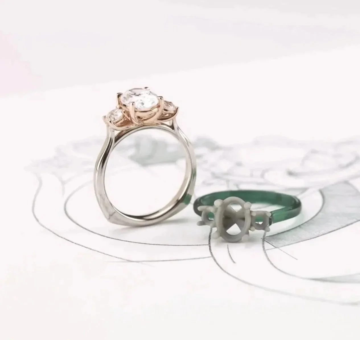 A diamond engagement ring with a rose gold band and a smaller ring with a green band and nautical wheel design, both placed on a sketch of a wedding dress.
