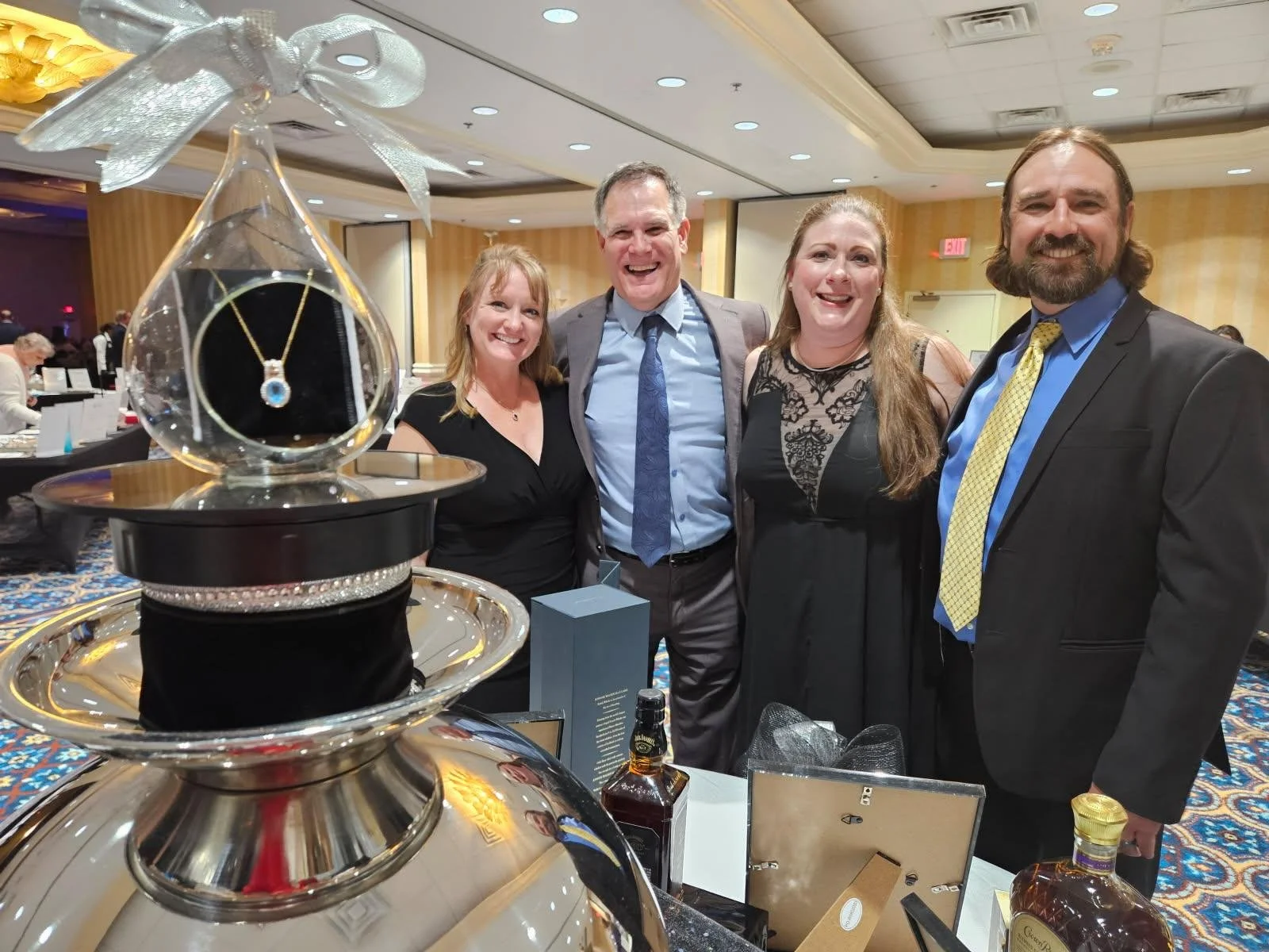 Four people, two women and two men, standing together at a formal event, smiling for the camera, with jewelry displayed in the foreground.