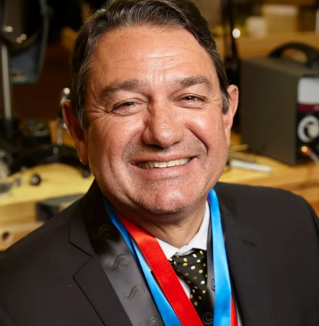 Smiling man in a suit with medals around his neck, in a setting with sound equipment and woodworking tools in the background.
