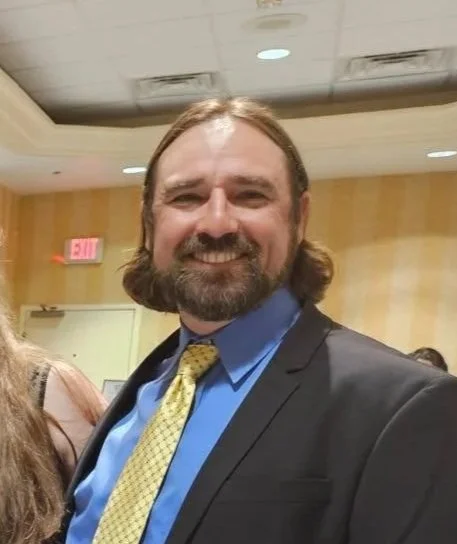 Smiling man with shoulder-length hair and a beard, wearing a suit and yellow tie, at an indoor event.
