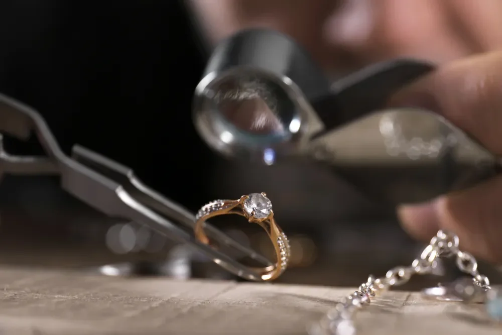 Close-up of hand holding a jeweler's loupe examining an engagement ring with a large central diamond, on a table with jewelry and tools.
