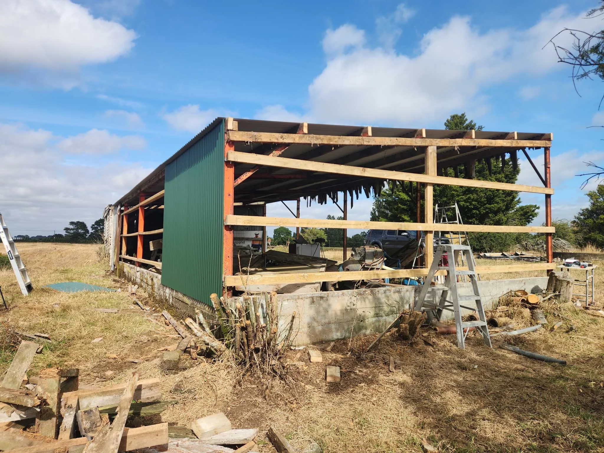 Construction site with an unfinished building frame, ladders, tools, and green siding, set in a rural area with trees and a blue sky.