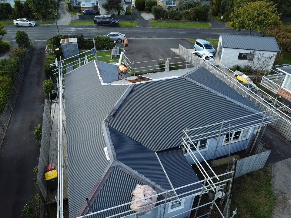 A house under construction with workers installing a new metal roof, surrounded by scaffolding, fences, and neighboring houses and cars in a residential area.