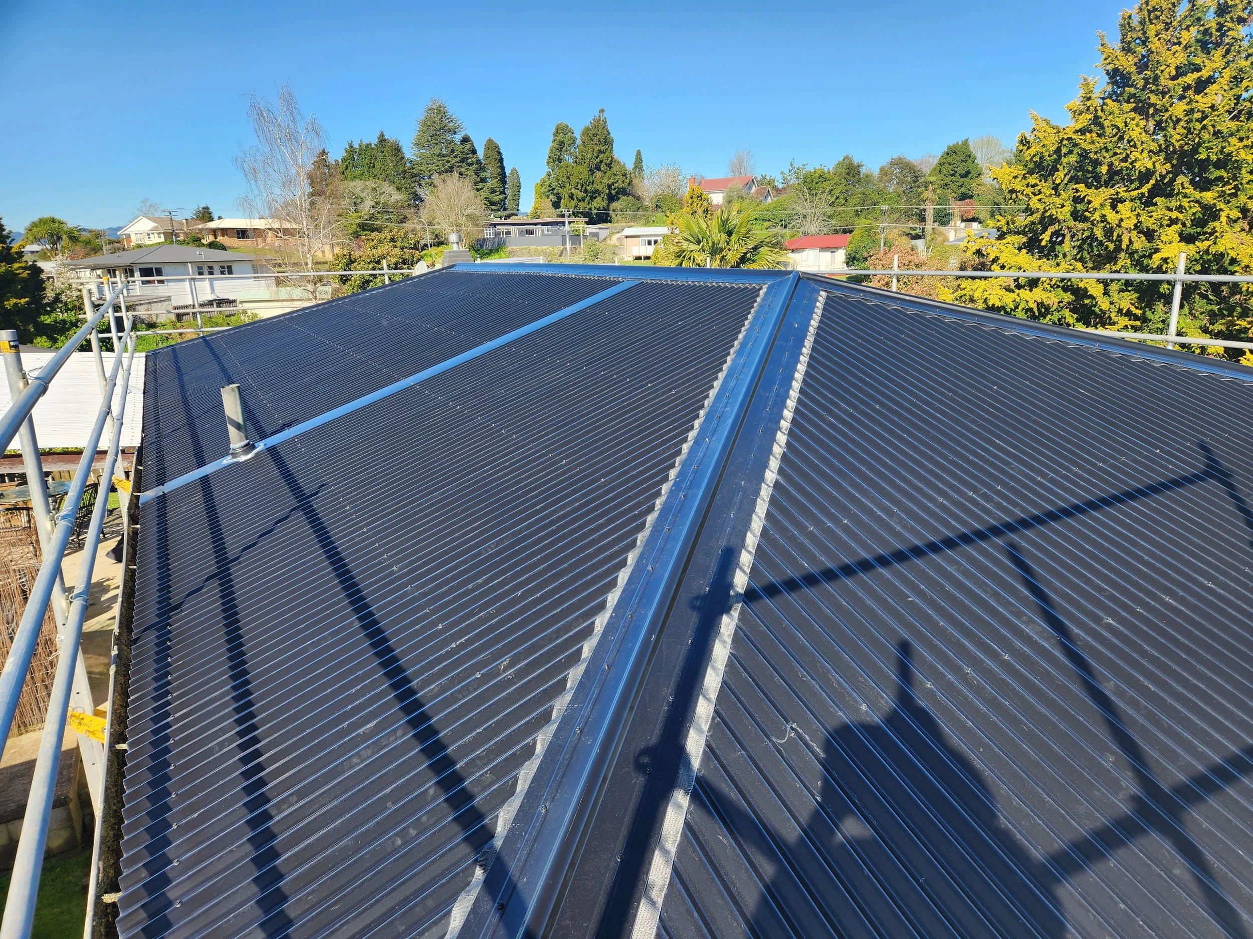 View of a rooftop covered with black solar panels, surrounded by a safety railing, with trees and houses in the background under a clear blue sky.