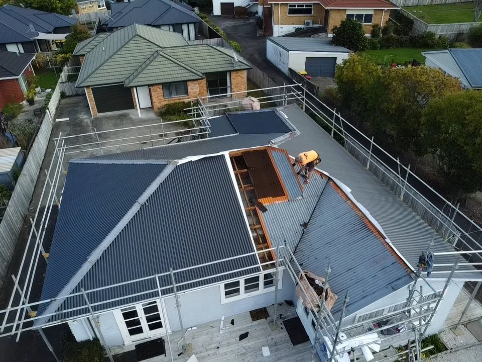 A construction worker on the roof of a house replacing or repairing the metal roofing panels. The house is surrounded by scaffolding, and there are neighboring houses and trees in the background.
