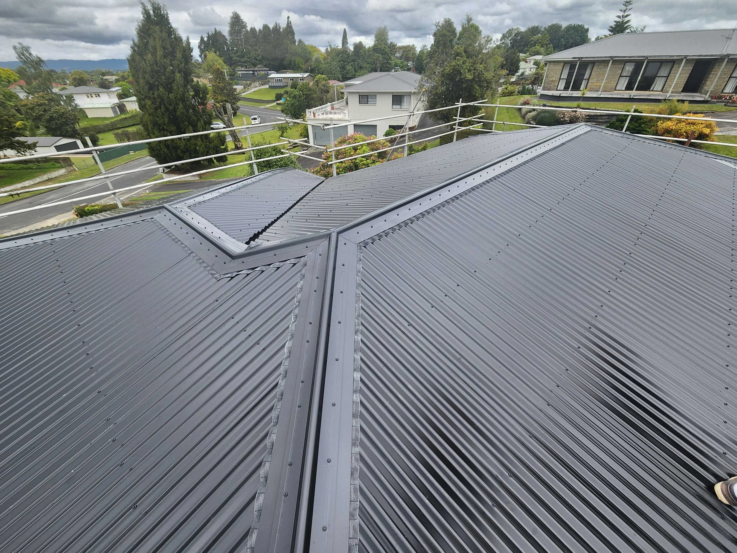 View of a rooftop with gray metal panels surrounded by safety railings, with a neighborhood of houses, trees, and a cloudy sky in the background.