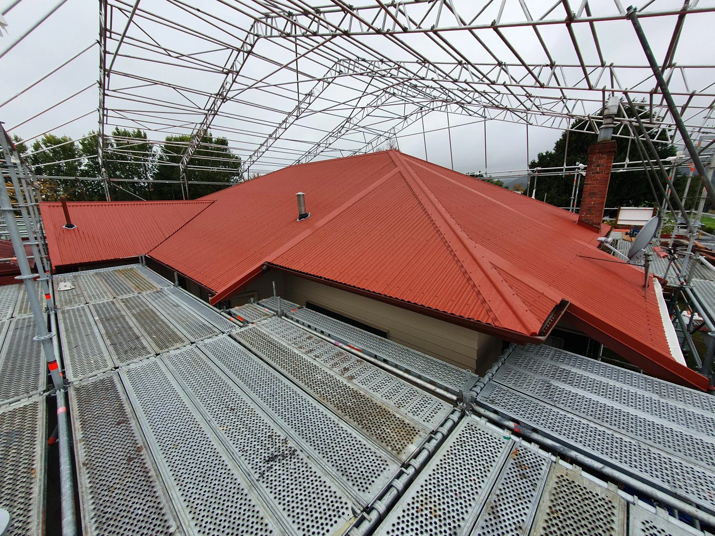 Construction scaffolding surrounding a house with a red metal roof and chimney, under an overcast sky.