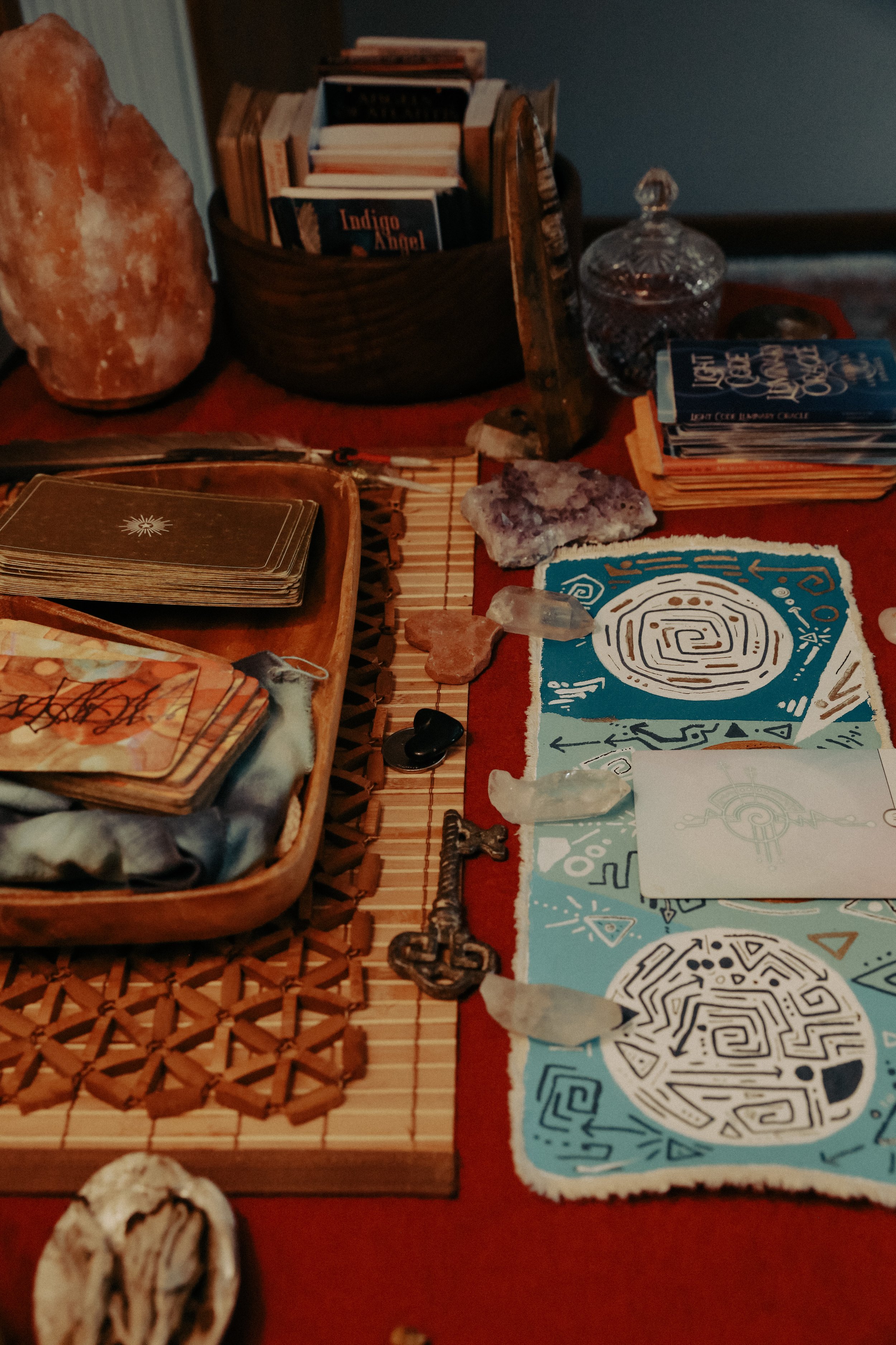 A table with tarot cards, crystals, and artwork on a red tablecloth, along with rocks and a box of playing cards.