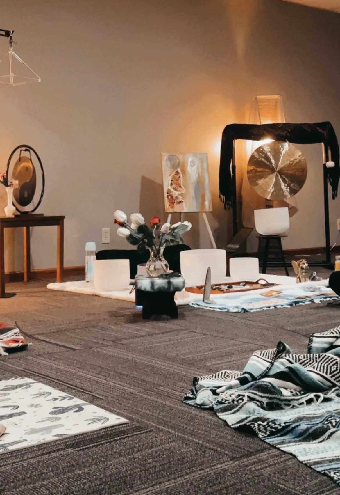 Sound Healing Omaha. Indoor space with meditation or yoga setup including crystal bowls, a vase of white flowers, and a tiger figurine, with various wall art and decor items like paintings, a gong, and a black curtain.