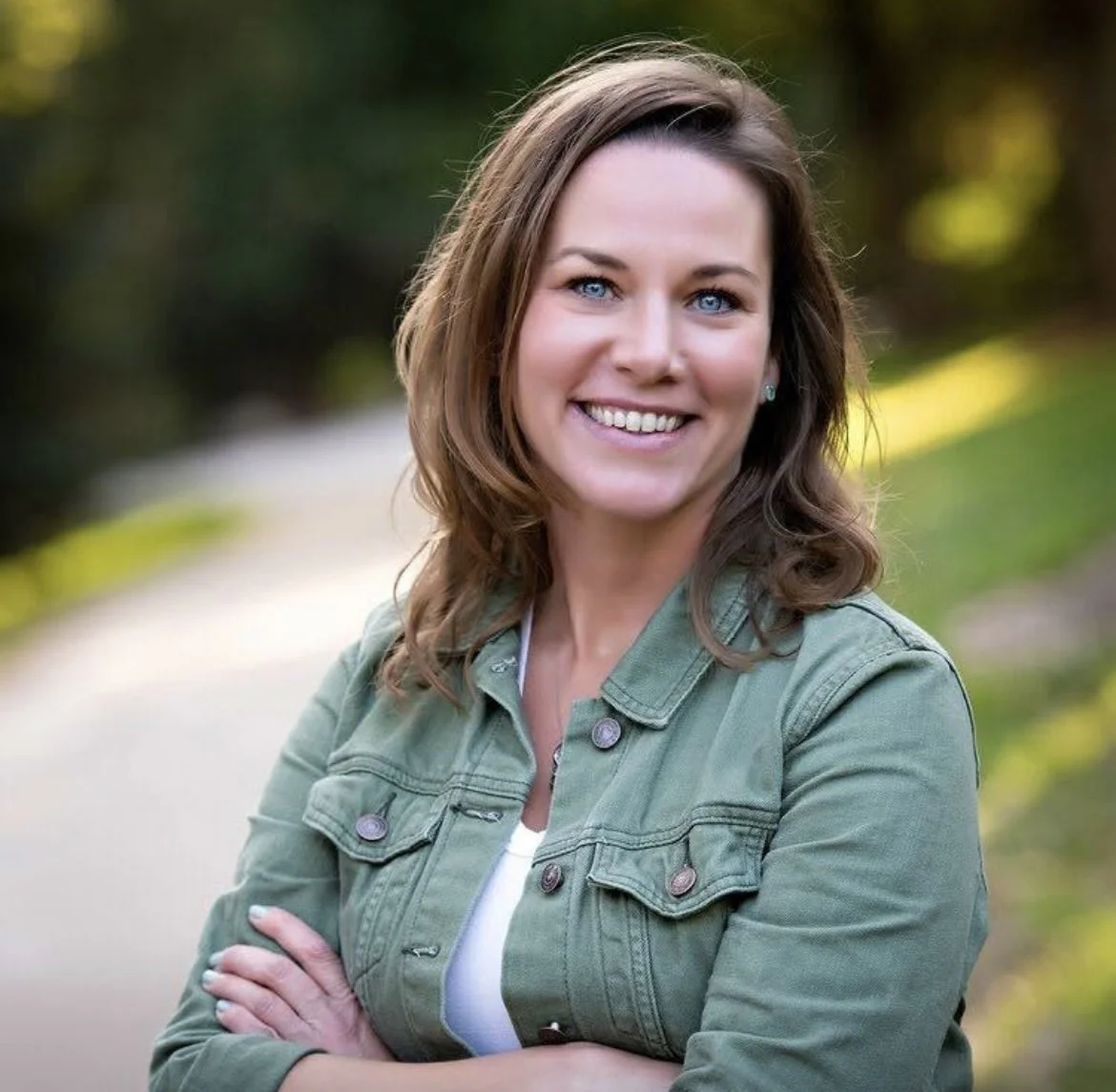 A woman with light brown, wavy hair and blue eyes smiling outdoors, wearing a green jacket and a white shirt, with a blurred green and yellow background.