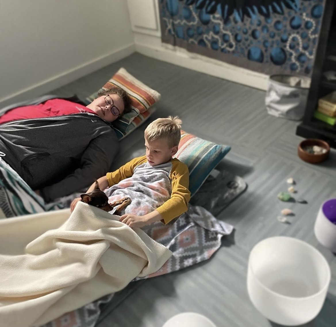 A young girl and a boy are lying on beds on the floor, with pillows under their heads. The girl appears to be sleeping, while the boy is awake, petting a small black and brown dog lying on his lap. They are in a room with a gray floor, a patterned cu