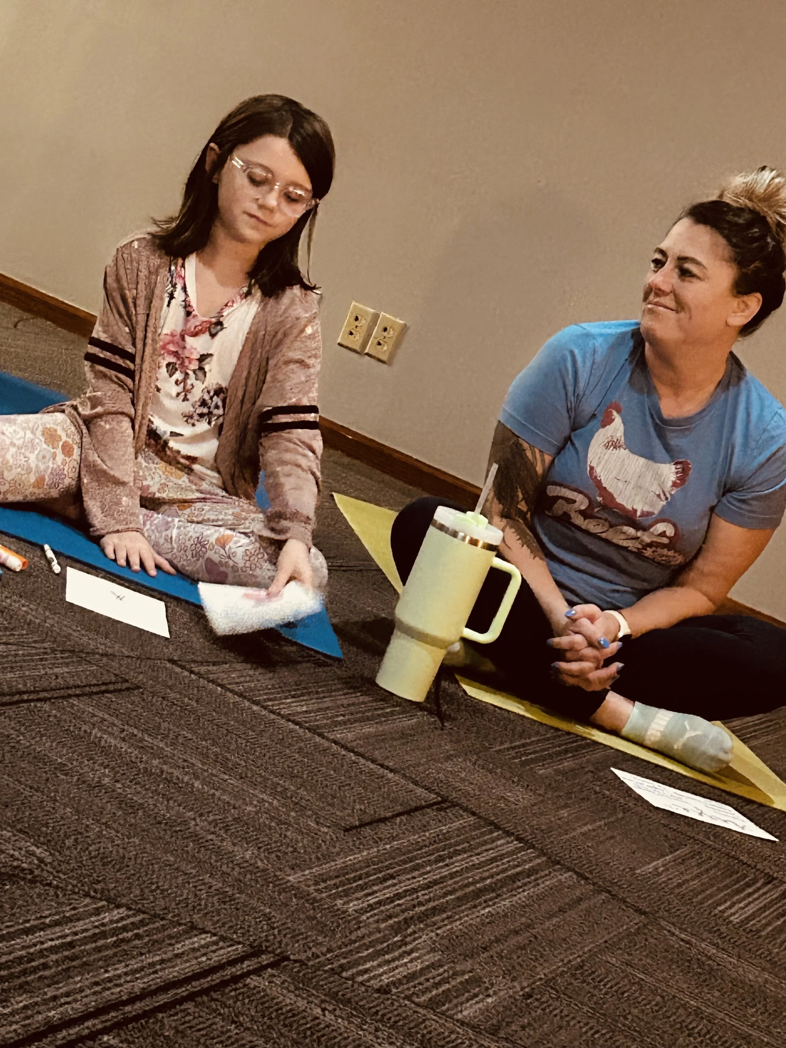 A woman in pajamas sitting on a yoga mat, reading a book, with a man beside her smiling. They are sitting on a carpeted floor in a room with beige walls and a yellow table.