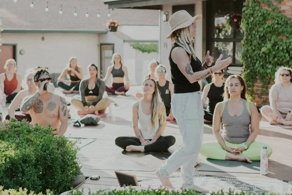 A woman in a hat leading a yoga class outdoors, with a group of women sitting cross-legged on mats, listening attentively.