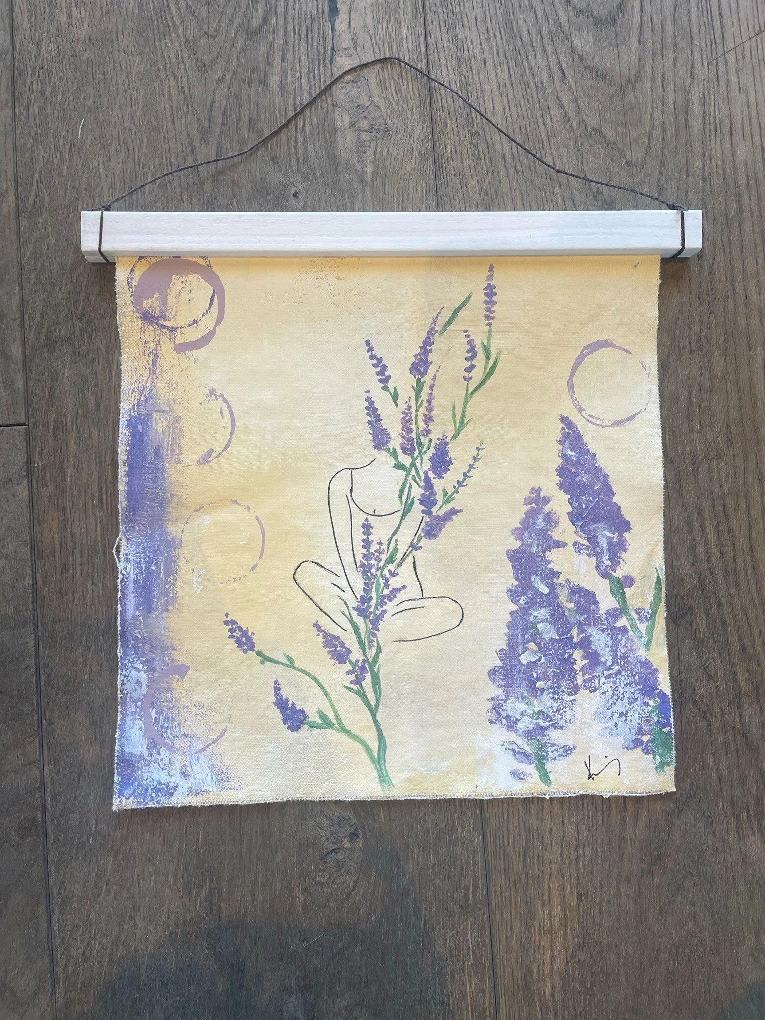 A paper hanging scroll featuring a drawing of a hand holding a bunch of lavender flowers, with purple circles and a purple lavender plant in the background, signed with initials.